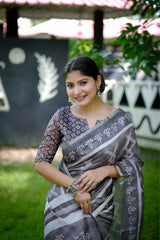 A woman models a striking Charcoal Grey Soft Cotton Saree featuring an all-over digital block print of geometric and abstract motifs in white. She wears a printed grey blouse and stacked silver bangles, posing outdoors against a stylized black and white wall.