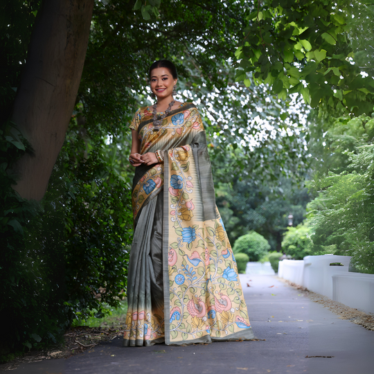 A woman models a sophisticated Slate Grey Soft Tussar Silk Saree. The saree features a wide pallu with a contrasting light yellow Kalamkari-style print, decorated with large floral motifs in dusty pink, blue, and green. She wears a matching short-sleeve printed blouse and accessorizes with heavy oxidized silver jewelry, posing outdoors.