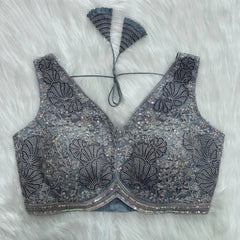 Elegant grey blouse featuring intricate sequin and thread embroidery with floral and leaf motifs, deep V-neck with structured cups, geometric border, and tasseled tie-back detail, displayed flat on a white fur background.