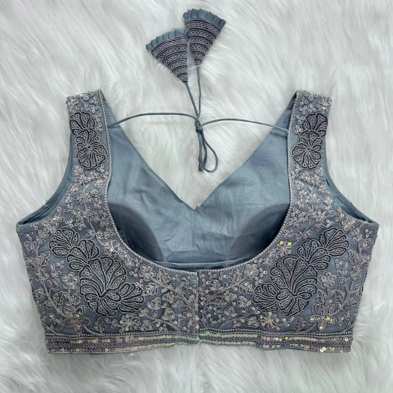 Elegant grey blouse featuring intricate sequin and thread embroidery with floral and leaf motifs, deep V-neck with structured cups, geometric border, and tasseled tie-back detail, displayed flat on a white fur background.