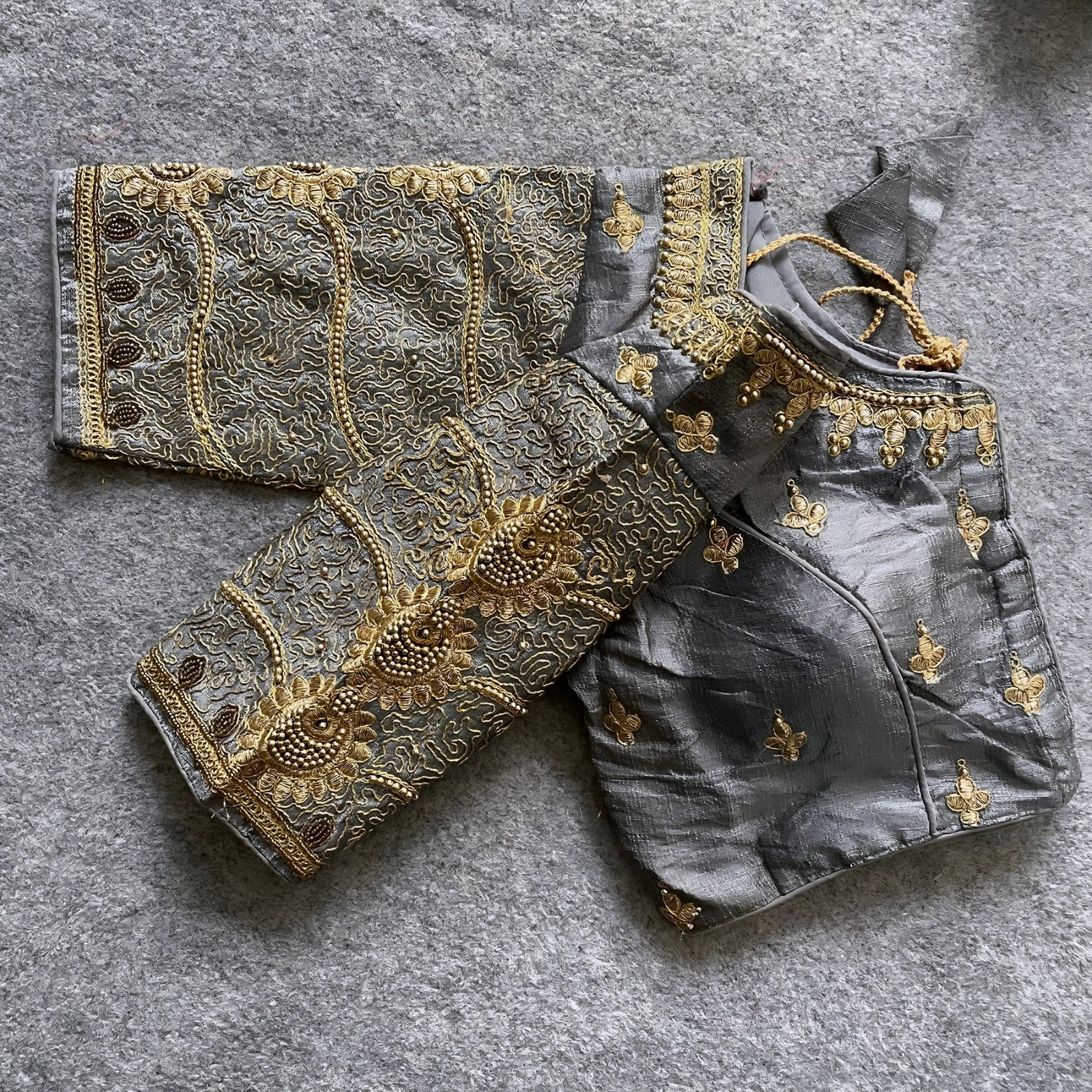 Traditional grey silk blouse featuring intricate gold embroidery and beadwork with floral and paisley motifs on the sleeves and neckline, displayed flat on a textured surface with a bunch of artificial flowers and a woven mat in the background.