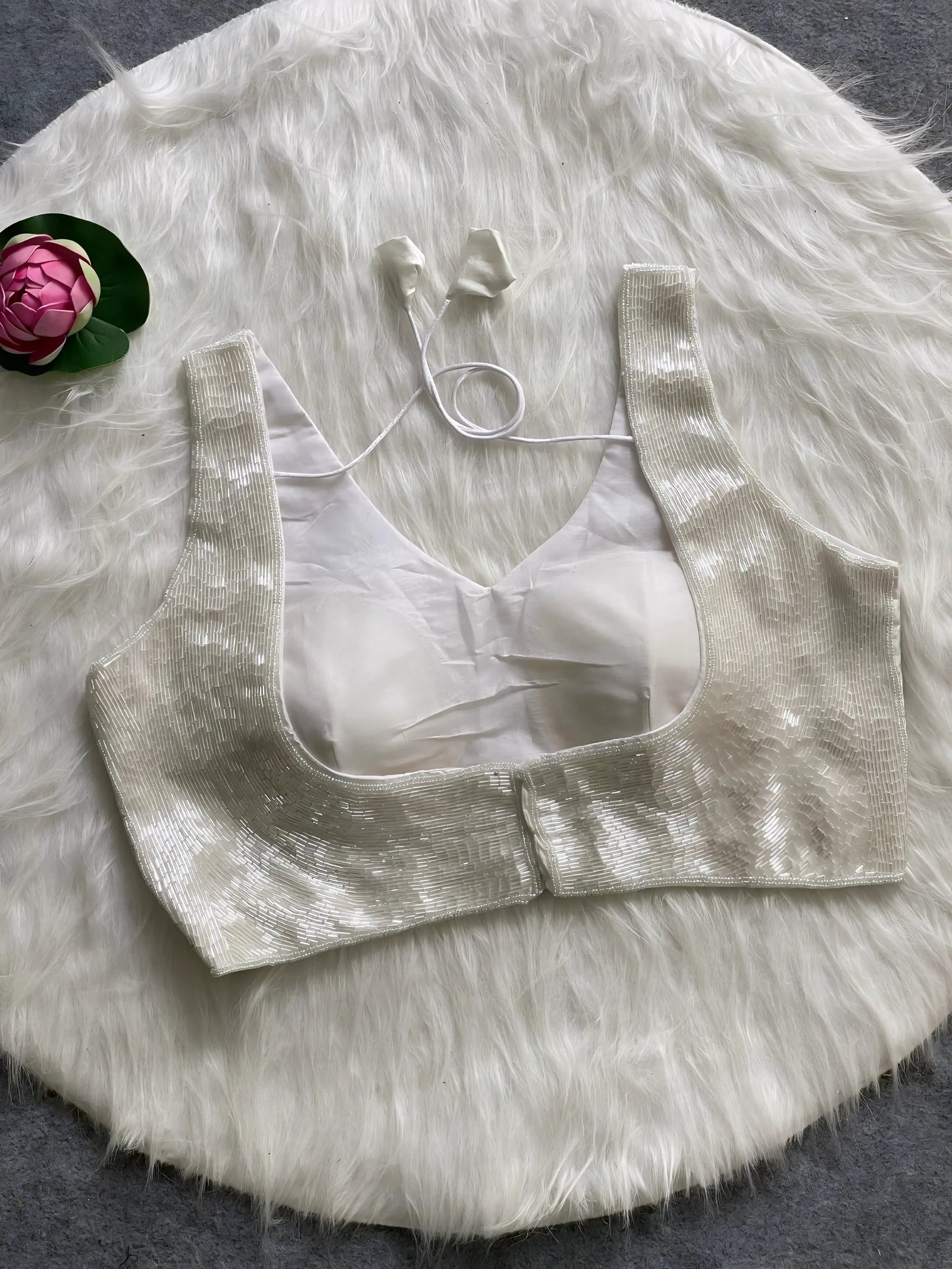 Ivory sequin sleeveless blouse with tie-back detail, displayed on a white fur background with a pink floral prop on the side