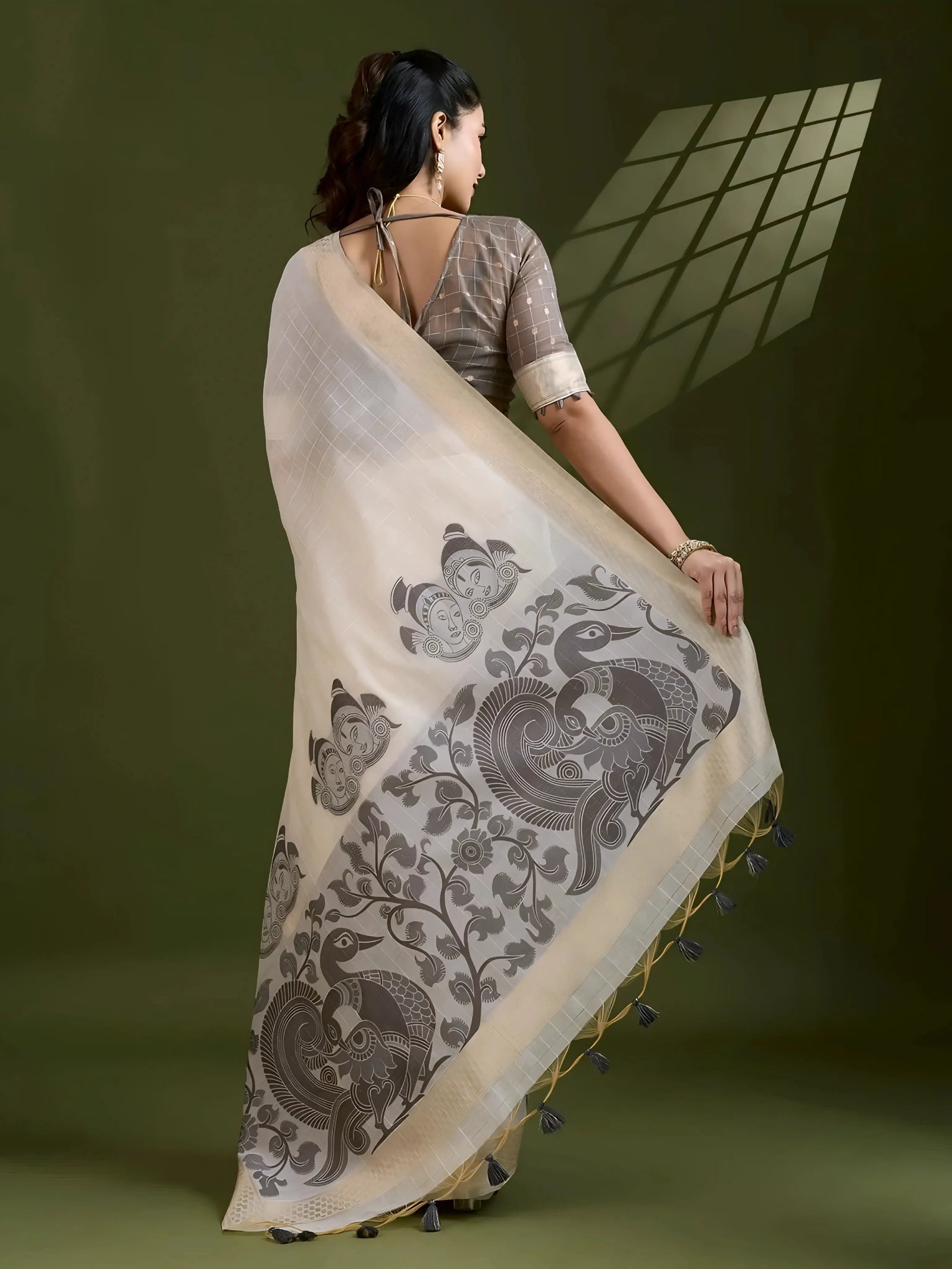 A woman models an elegant Beige Soft Muga Cotton Saree. The saree features large Kalamkari-style prints of peacocks, trees, and florals in a contrasting Dark Gray on the pallu. She wears a dark gray short-sleeve sheer blouse and accessorizes with a gold Kundan choker necklace and sunglasses.