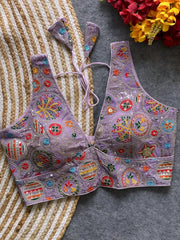 Traditional lavender blouse featuring vibrant multicolor thread embroidery and mirror work in circular floral and geometric patterns, with sequin accents and a deep V-neck tie-back detail, displayed flat on a textured mat with artificial red and yellow flowers in the corner.
