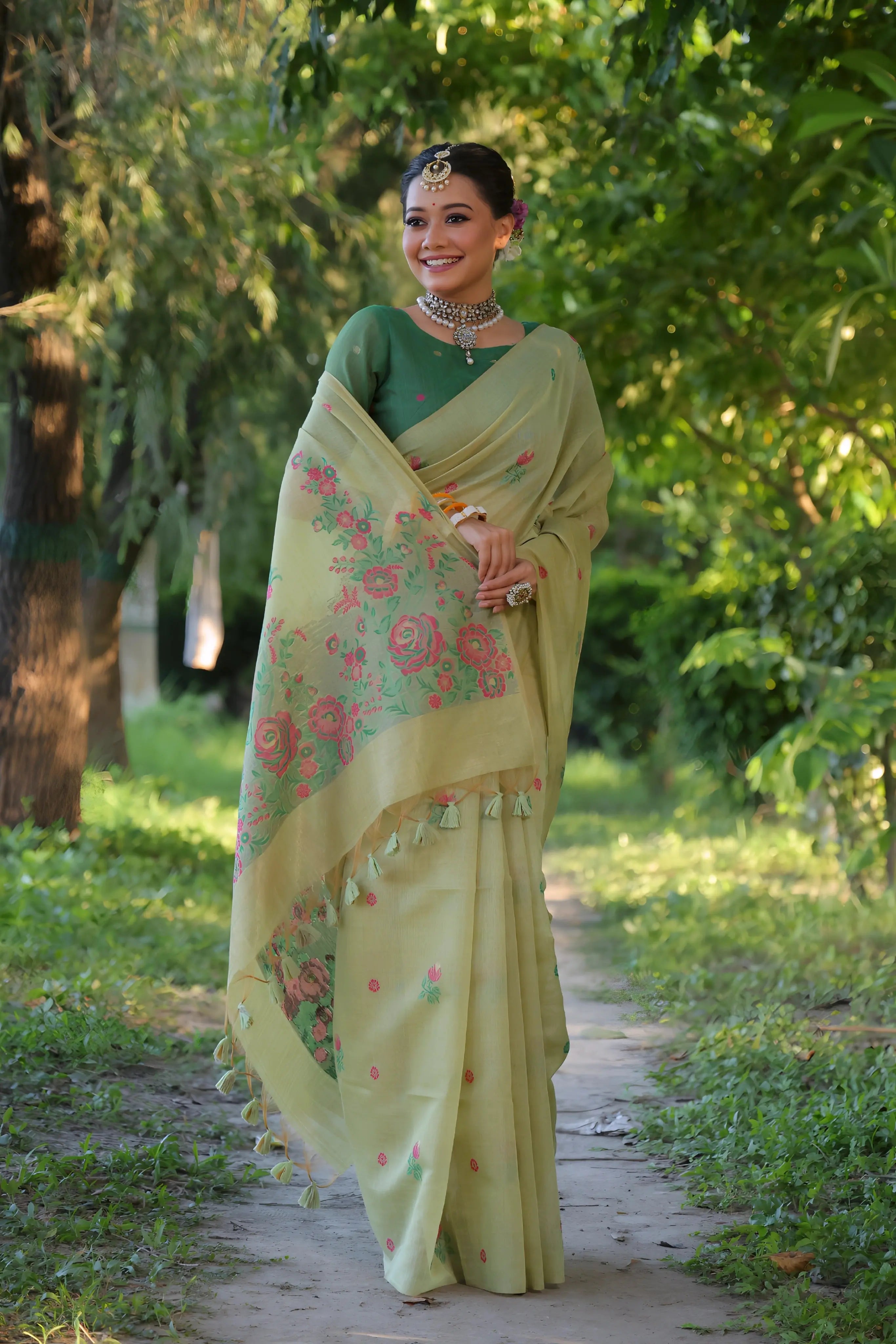 A woman models an elegant light Pista Green Soft Muga Cotton Saree with scattered pink and red floral Resham weaving. The pallu features an elaborate panel of multi-colored floral weaving/print. She wears a contrasting deep green blouse, white stone/Kundan jewelry, and white and orange bangles, posing outdoors.