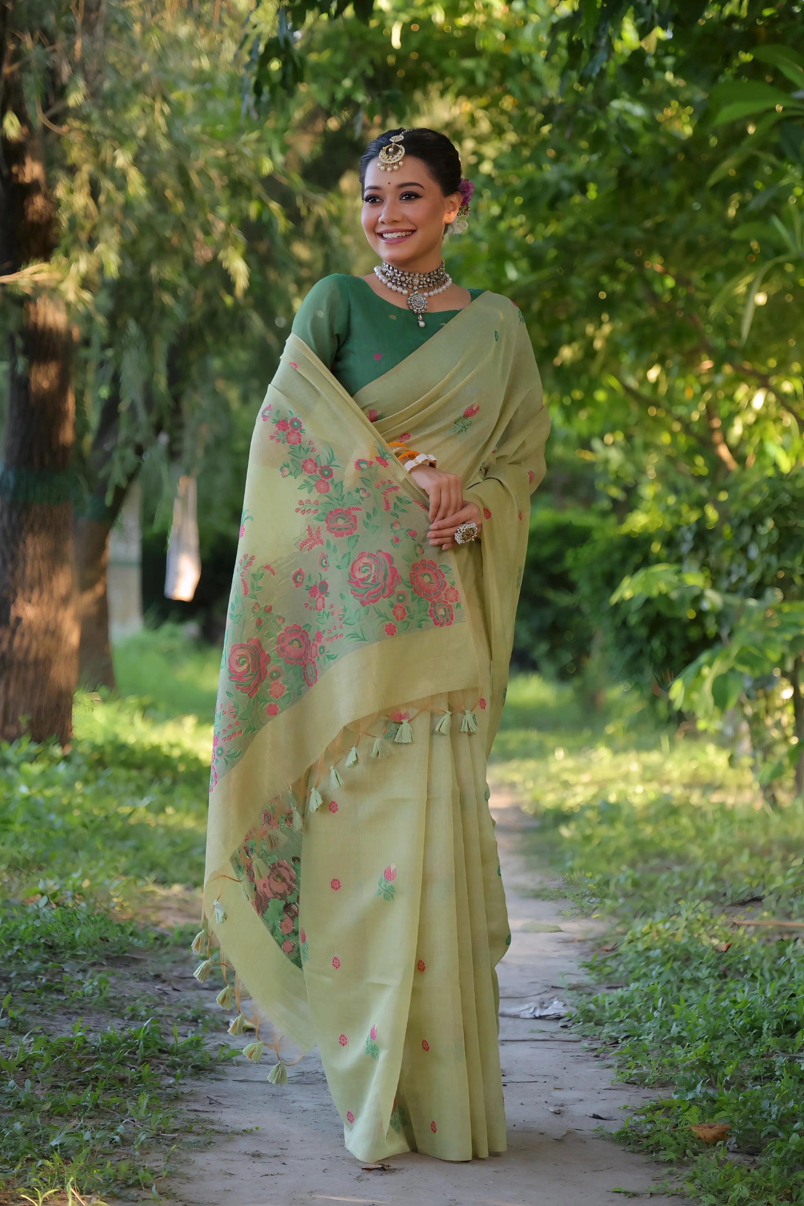 A woman models an elegant light Pista Green Soft Muga Cotton Saree with scattered pink and red floral Resham weaving. The pallu features an elaborate panel of multi-colored floral weaving/print. She wears a contrasting deep green blouse, white stone/Kundan jewelry, and white and orange bangles, posing outdoors.