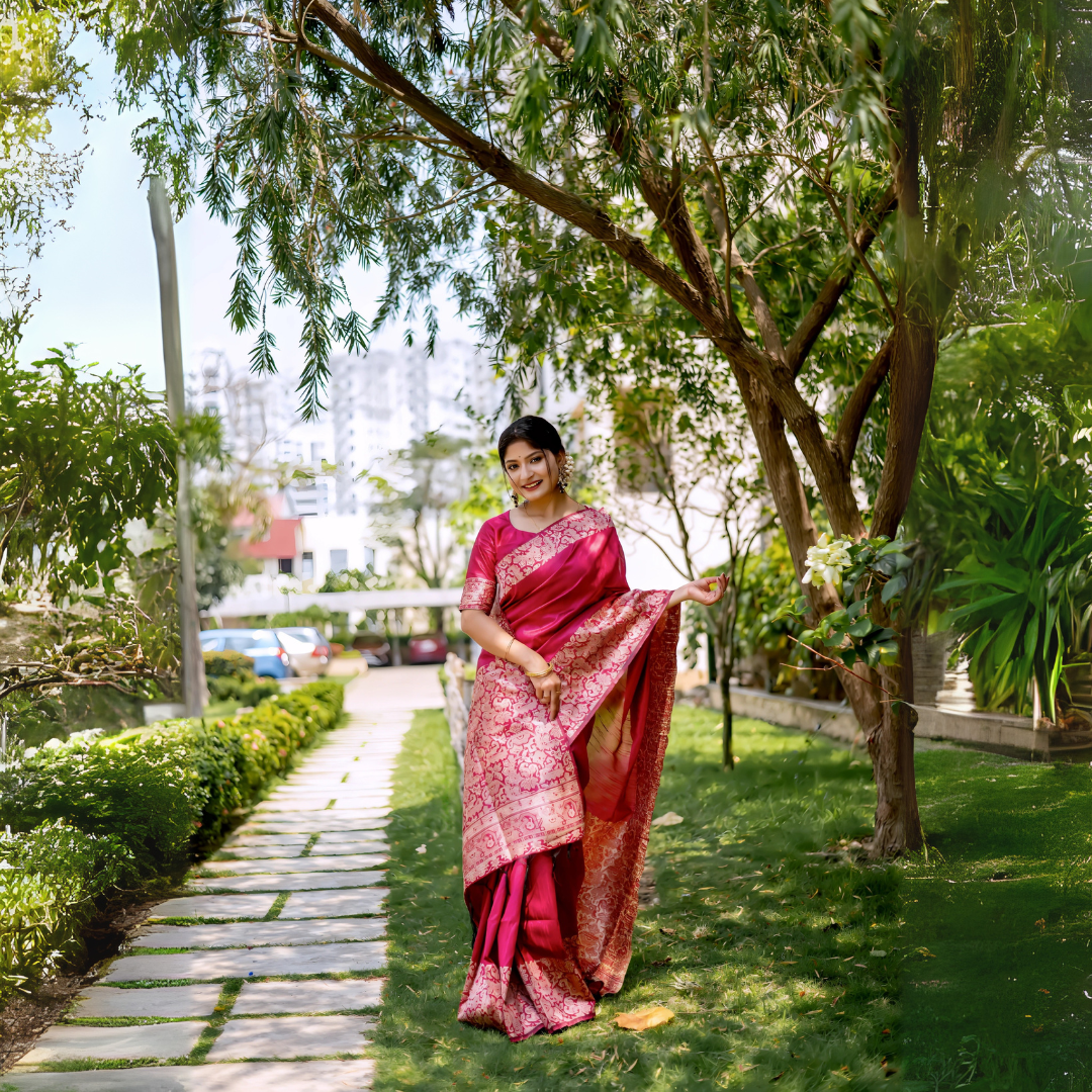 A woman models a luxurious Deep Pink Raw Silk Saree. The saree features a wide border and pallu with dense, intricate gold Zari weaving in a Kalamkari-style floral and paisley pattern. She wears a matching short-sleeve pink blouse and traditional gold jewelry, posing outdoors on a stone pathway, framed by lush greenery.
