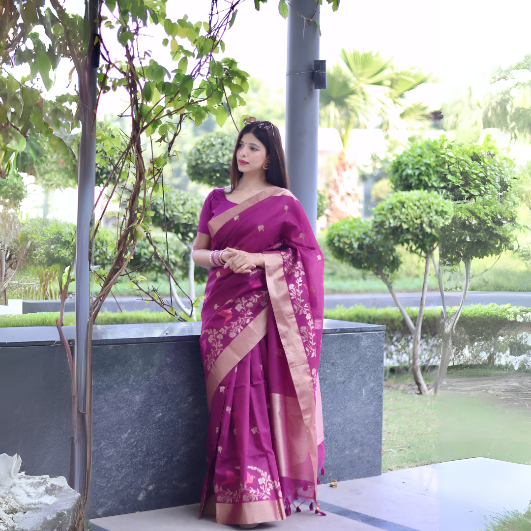 A woman models a vibrant Fuchsia Purple Linen Silk Saree with delicate gold Zari woven floral motifs. The saree has a wide metallic gold border. She wears a matching purple blouse and subtle gold jewelry, posing in a modern outdoor setting.