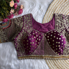 Traditional magenta silk blouse featuring intricate gold zardosi and thread embroidery with floral and peacock motifs, sequin embellishments, geometric panel designs, and detailed sleeves, displayed flat on a white fur surface with a braided mat and artificial pink flowers in the corner.