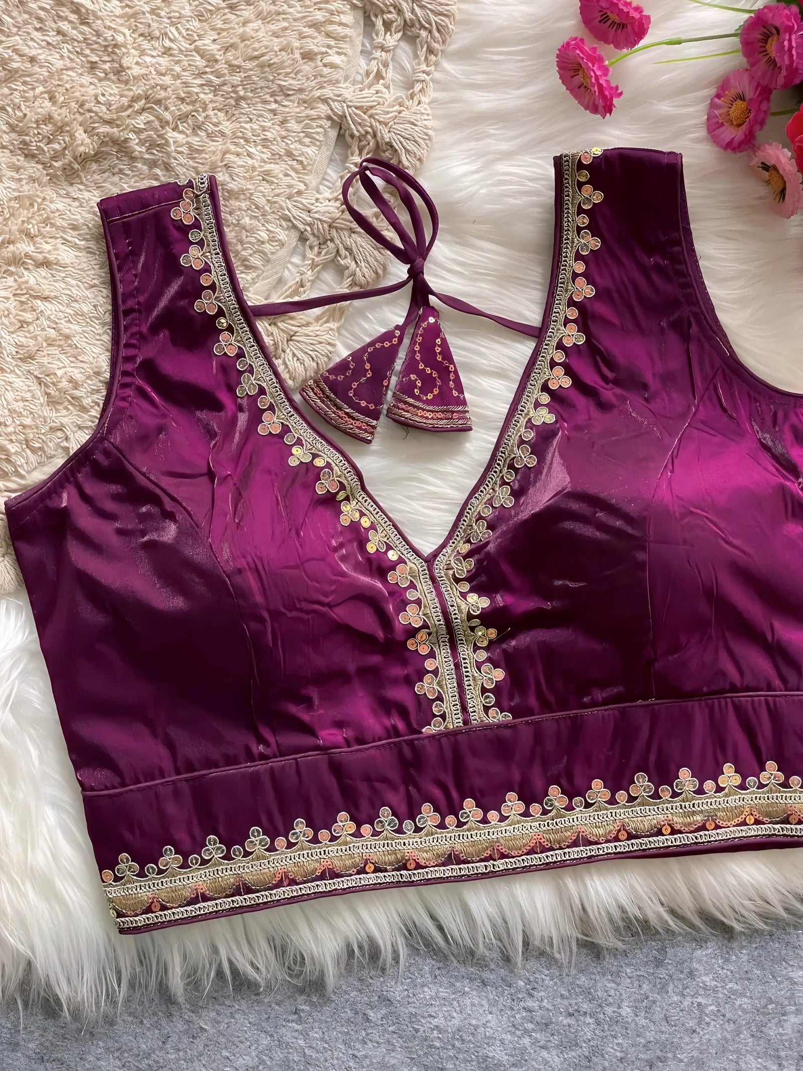 Traditional magenta silk blouse featuring intricate gold embroidery and beadwork with floral motifs along the neckline and hem, displayed flat on a soft fur and textured mat background with pink artificial flowers in the corner.