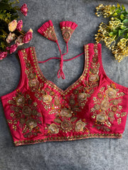 Traditional magenta silk blouse featuring intricate gold zardozi embroidery and beadwork with floral and leaf motifs on the front and neckline, finished with decorative tassel ties, displayed flat on a textured grey background with artificial pink and yellow flowers in the corners.