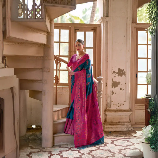 Woman wearing a magenta silk saree with teal pallu and intricate floral motifs, paired with matching blouse and traditional earrings, elegantly posing beside a vintage spiral staircase in a sunlit heritage interior with large wooden windows, distressed textured walls, and geometric patterned tile flooring.