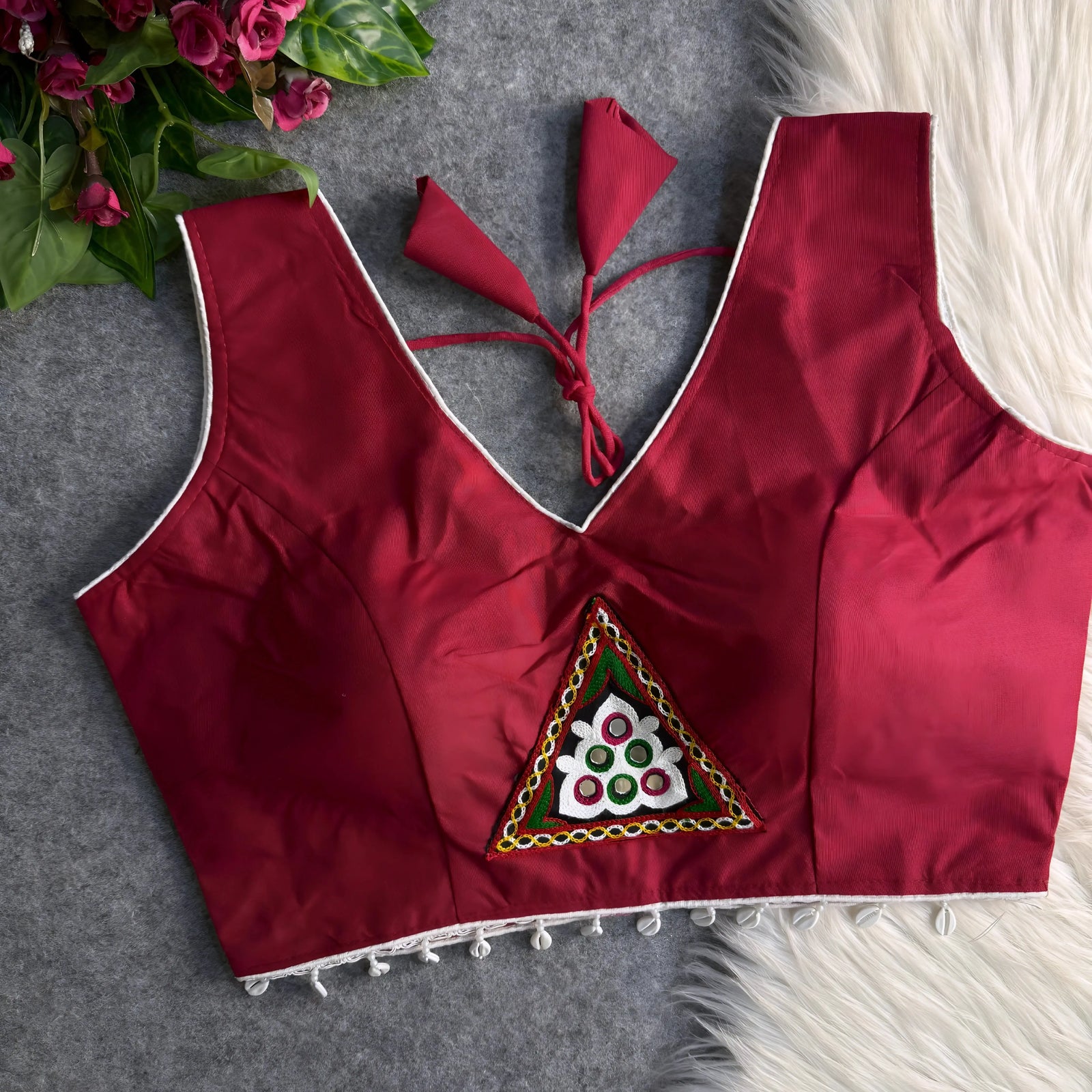 Maroon padded blouse with white piping, featuring a central embroidered triangle motif with mirror work and shell detailing along the hem, displayed on a grey surface with artificial pink flowers and white fur in the background.