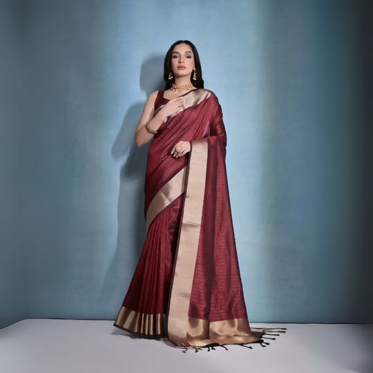 A woman models a striking Dark Maroon Banarasi Raw Silk Saree. The saree features a very wide, luxurious metallic gold Zari border and subtle woven stripes on the body. She wears a sleeveless maroon blouse and traditional gold Kundan jewelry, posing indoors.