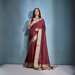 A woman models a striking Dark Maroon Banarasi Raw Silk Saree. The saree features a very wide, luxurious metallic gold Zari border and subtle woven stripes on the body. She wears a sleeveless maroon blouse and traditional gold Kundan jewelry, posing indoors.