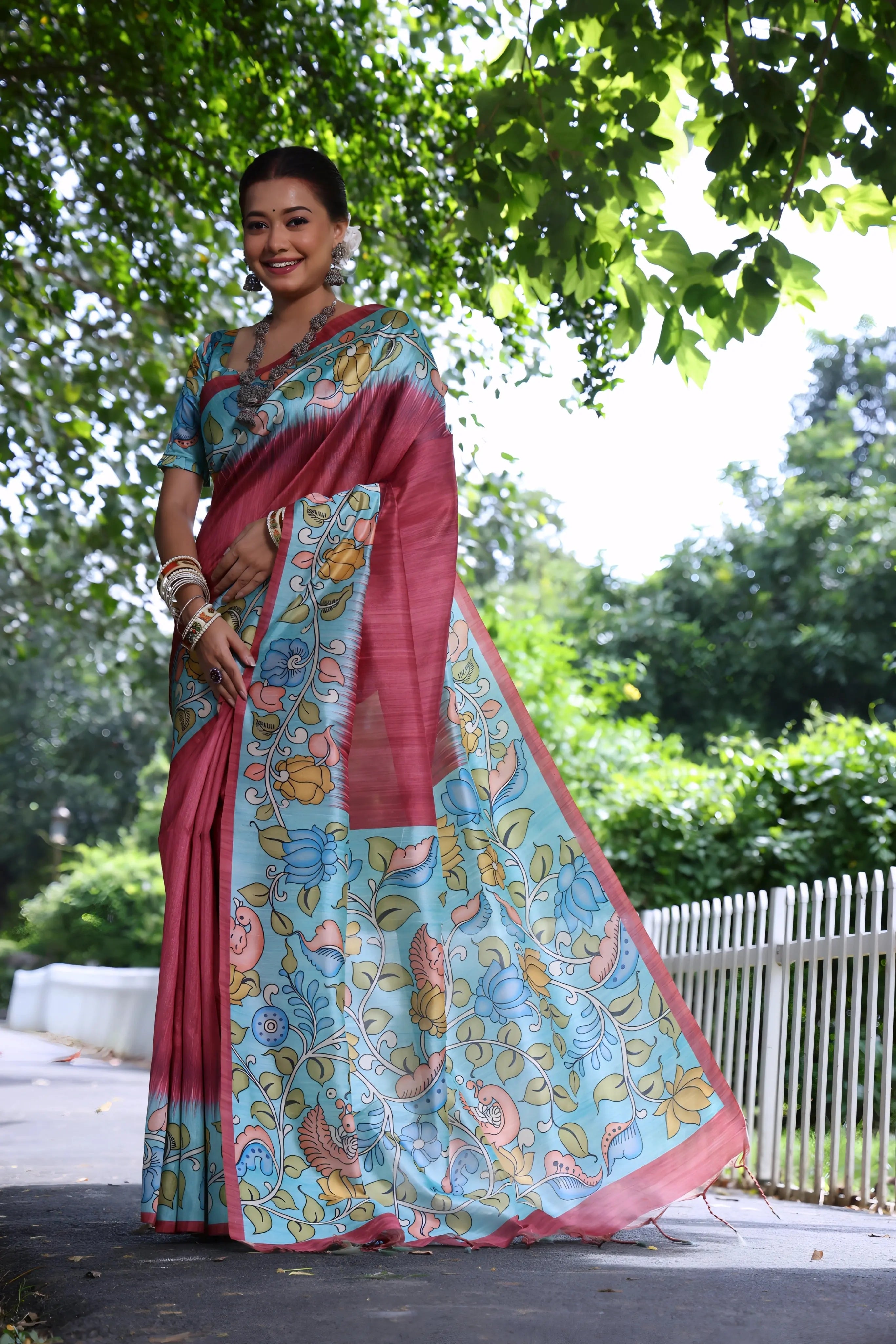 A woman models a stunning Rose Red Soft Tussar Silk Saree. The wide border and pallu feature an elaborate Kalamkari-style print on a Sky Blue base, with large bird and floral motifs in soft pink, yellow, and blue. She wears a matching printed blouse and accessorizes with heavy oxidized silver jewelry, posing outdoors in a garden.