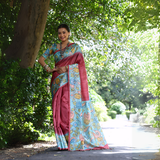 A woman models a stunning Rose Red Soft Tussar Silk Saree. The wide border and pallu feature an elaborate Kalamkari-style print on a Sky Blue base, with large bird and floral motifs in soft pink, yellow, and blue. She wears a matching printed blouse and accessorizes with heavy oxidized silver jewelry, posing outdoors in a garden.