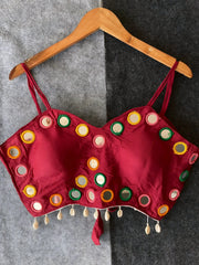 Maroon corset-style blouse adorned with vibrant round mirrors and multicolored embroidered borders, featuring thin shoulder straps and shell tassel drops along the hem. Displayed on a wooden hanger against a two-tone grey textured background, highlighting its festive and contemporary ethnic appeal