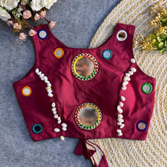 Deep maroon ethnic blouse with colorful circular mirror work and cowrie shell detailing arranged on a woven mat.