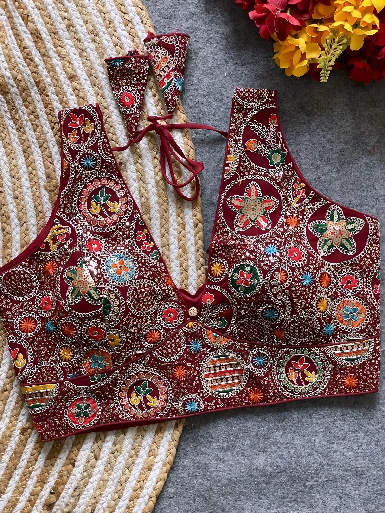 Traditional maroon blouse featuring vibrant multicolor thread embroidery and mirror work in circular floral and geometric patterns, with a deep V-neck and tie-back detail, displayed flat on a textured mat with artificial red and yellow flowers in the corner.