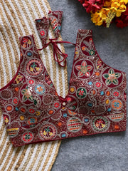 Traditional maroon blouse featuring vibrant multicolor thread embroidery and mirror work in circular floral and geometric patterns, with a deep V-neck and tie-back detail, displayed flat on a textured mat with artificial red and yellow flowers in the corner.