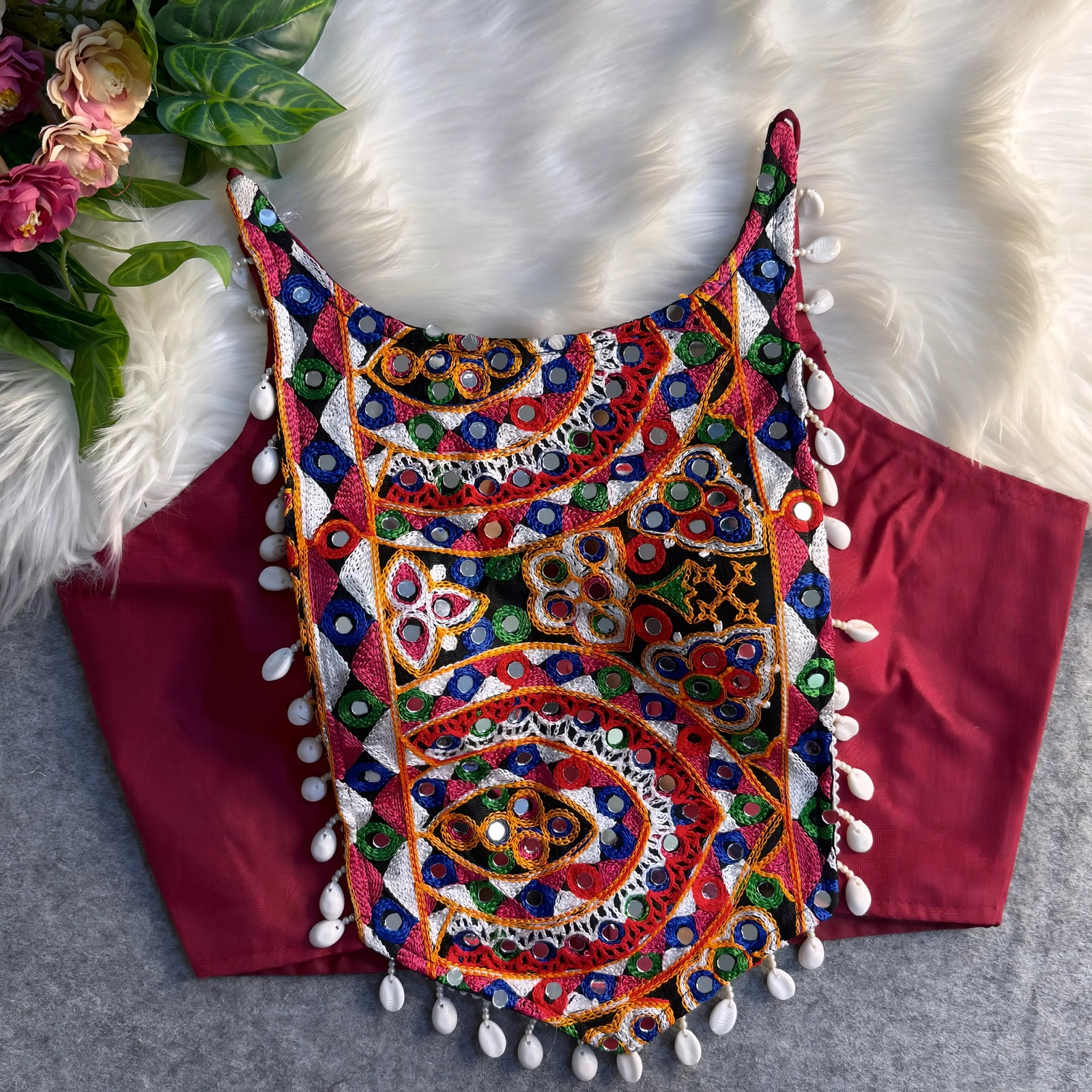 Traditional maroon blouse featuring vibrant multicolor mirror work and intricate thread embroidery in geometric and paisley patterns, accented with white shell tassels along the hem, displayed flat on a soft fur and grey background with floral and leafy decor.