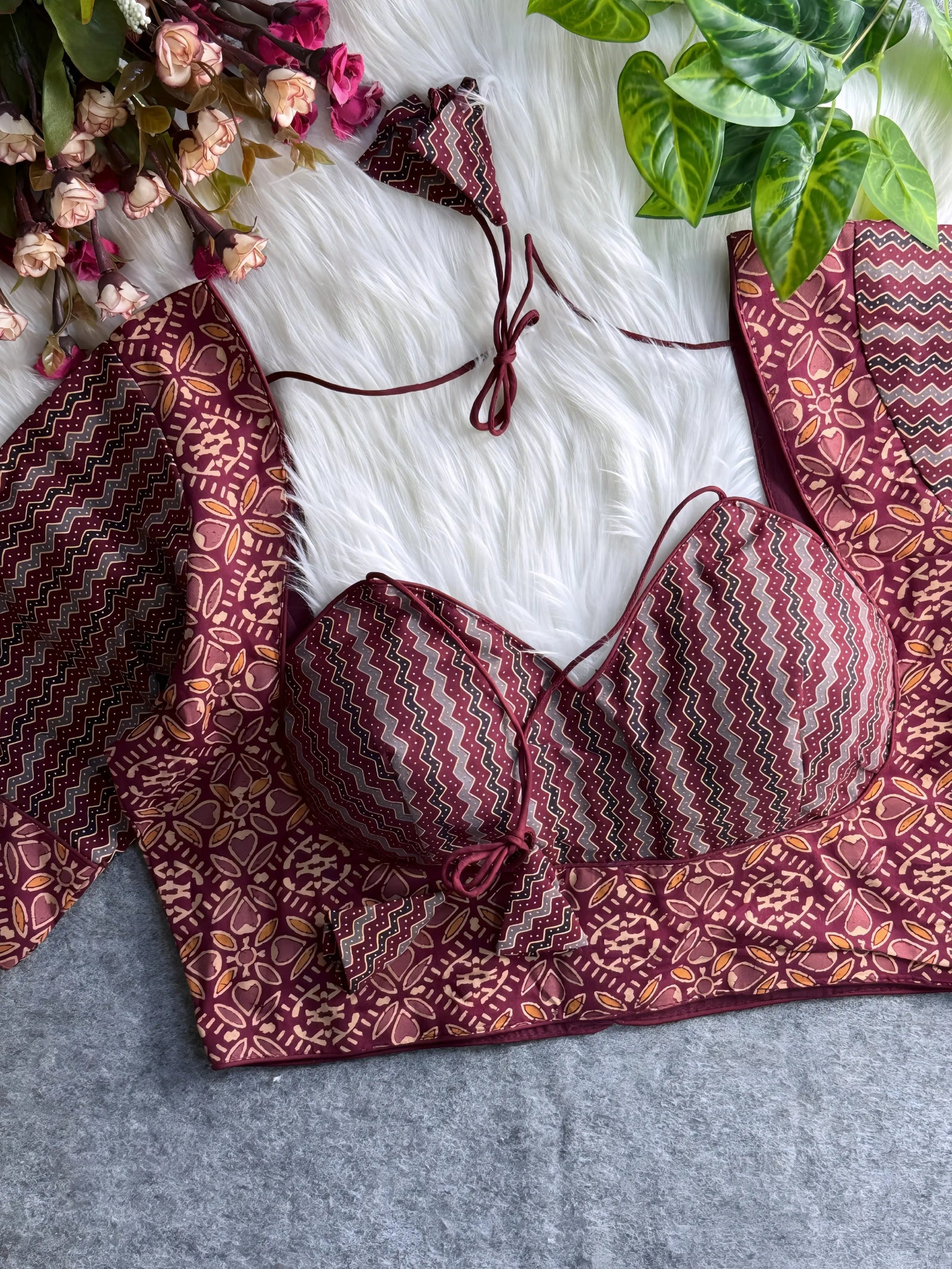 Maroon printed padded blouse with geometric and floral patterns, displayed on a white fur surface with artificial flowers and green leaves in the background.