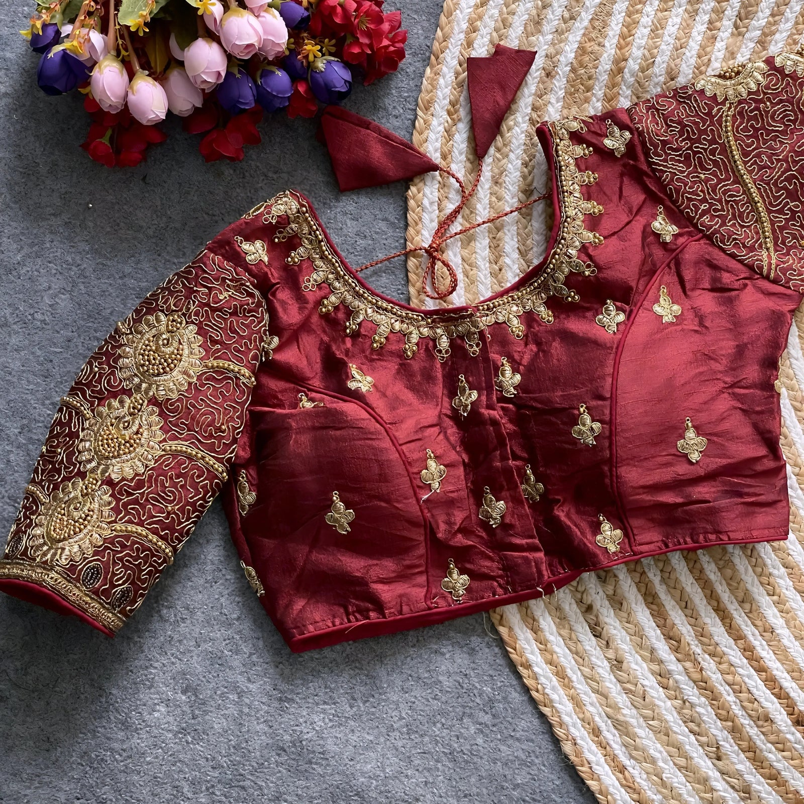 Traditional maroon silk blouse featuring intricate gold embroidery and beadwork with floral and paisley motifs on the sleeves and neckline, displayed flat on a textured surface with a bunch of artificial flowers and a woven mat in the background.