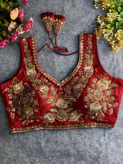 Traditional maroon silk blouse featuring intricate gold zardozi embroidery and beadwork with floral and leaf motifs on the front and neckline, finished with decorative tassel ties, displayed flat on a textured grey background with artificial pink and yellow flowers in the corners.