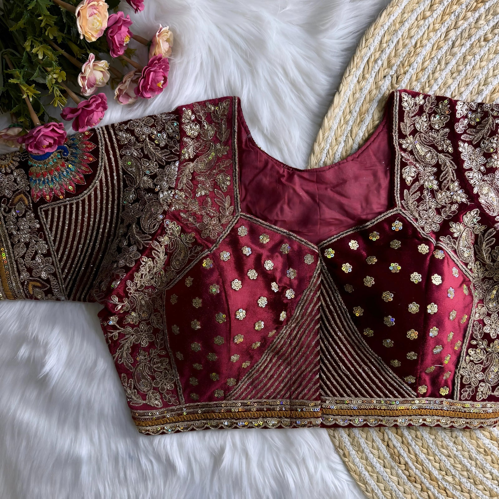 Maroon silk blouse featuring intricate gold zardosi embroidery with floral and paisley motifs, sequin polka dot accents, striped geometric panels, and a colorful peacock feather design on the sleeve, displayed flat on a white fur surface with a braided mat and pink artificial flowers in the corner.