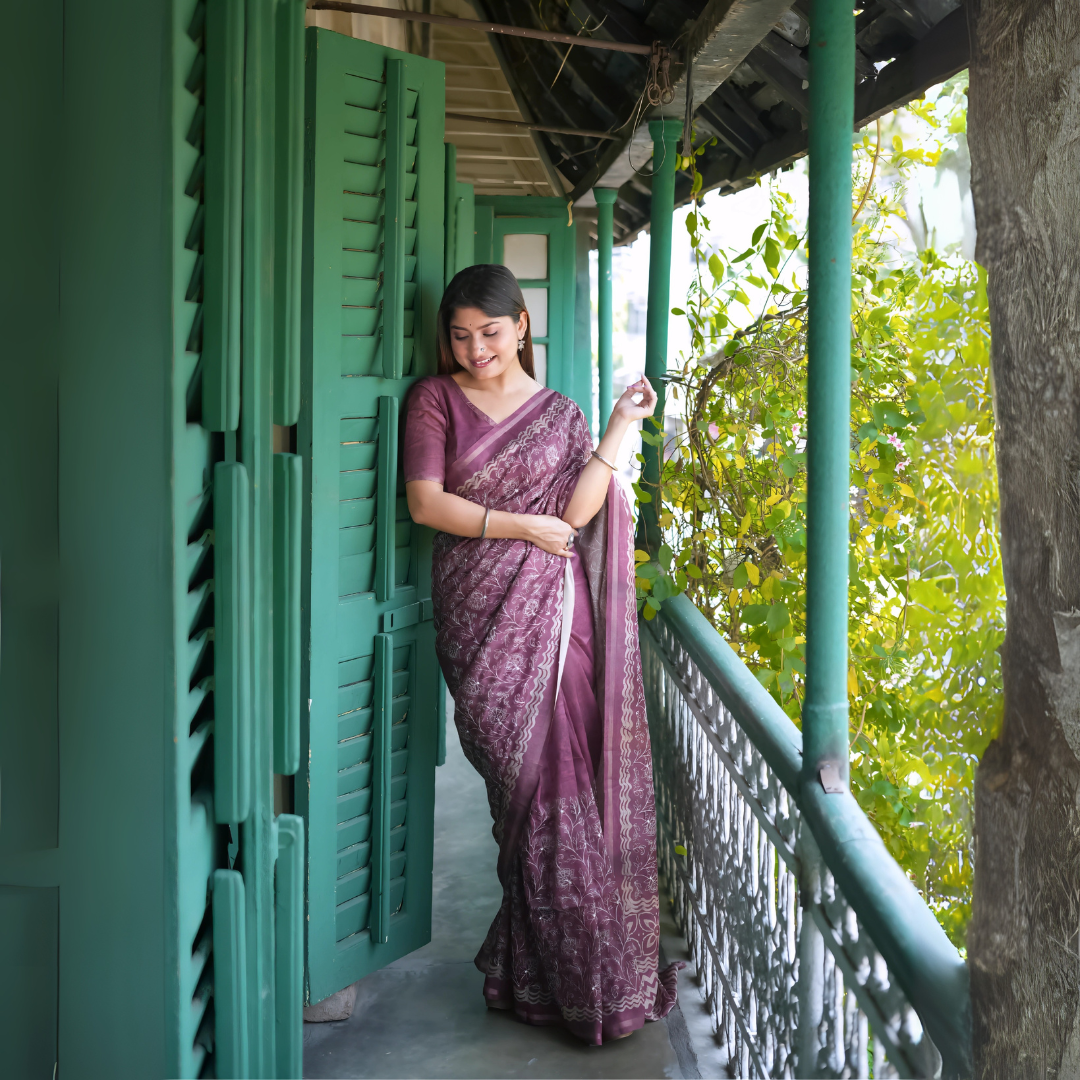 A woman models an elegant Wine Red/Deep Maroon Tussar Silk Saree. The saree features dense all-over floral embroidery/weaving in a light metallic color, framed by a contrasting light metallic border. She wears a matching wine short-sleeve blouse and subtle silver bangles, posing on a balcony framed by vibrant teal shutters.