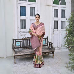 A woman models an elegant Light Peach Soft Silk Saree featuring a large-scale floral and figurative Pichwai-style print in soft colors. The saree has a wide Deep Brown/Maroon border and pallu. She wears a matching dark blouse and a gold Kundan choker necklace, posing outdoors next to distressed antique white doors.