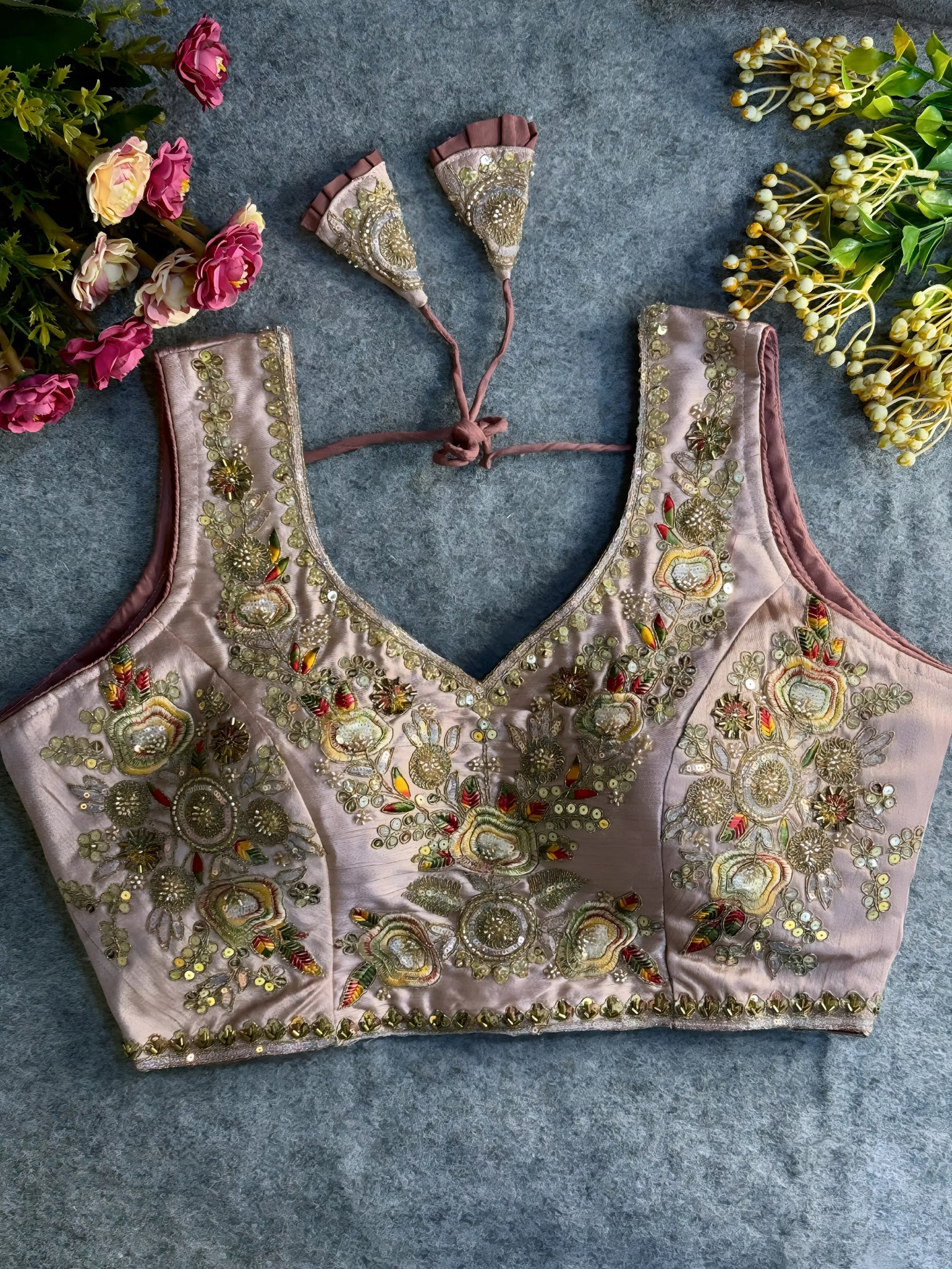 Traditional mauve silk blouse featuring intricate gold zardozi embroidery and beadwork with multicolor floral and leaf motifs on the front and neckline, finished with decorative tassel ties, displayed flat on a textured grey background with artificial pink and yellow flowers in the corners.