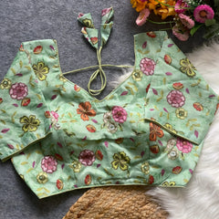 Mint green floral embroidered padded blouse with back tie detail, featuring multicolor threadwork flowers, displayed on a grey surface with artificial flowers and white fur in the background.