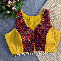Sleeveless yellow ethnic blouse showcasing vibrant mirror-work embroidery on the front panel, metallic chevron side panels, and shell tassels along the hem, laid on a grey surface with a woven mat and greenery accents