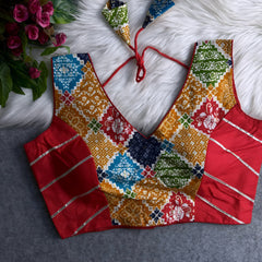Multicolor Ajrak-print blouse with deep V-neck, featuring traditional geometric and floral patterns in blue, mustard, green, gold, and red, contrasted by solid red silk side panels with silver stripes, and tie-back detail, displayed flat on a white fur background with a bunch of artificial pink flowers in the corner.