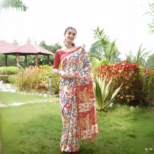 A woman models an elegant Beige Soft Tussar Silk Saree with a dense all-over print of multi-colored Kalamkari florals and vines. The saree has a wide, contrasting Deep Red border and pallu with traditional paisley print. She wears a matching red blouse and a chunky beaded necklace, posing outdoors in a bright garden setting.