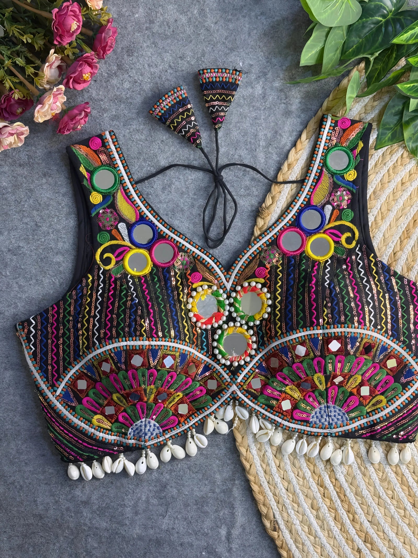 Traditional multicolor embroidered blouse featuring vibrant mirror work, intricate thread embroidery in geometric and floral patterns, and shell tassel detailing along the hem, displayed on a flat surface with floral and leafy decor.