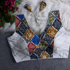 Traditional blouse featuring a multicolor patchwork pattern with intricate floral and geometric motifs, deep V-neckline, white silk side panels with gold accents, and patterned tasseled tie-back detail, displayed flat on a textured grey surface with artificial pink flowers and a white fur background.