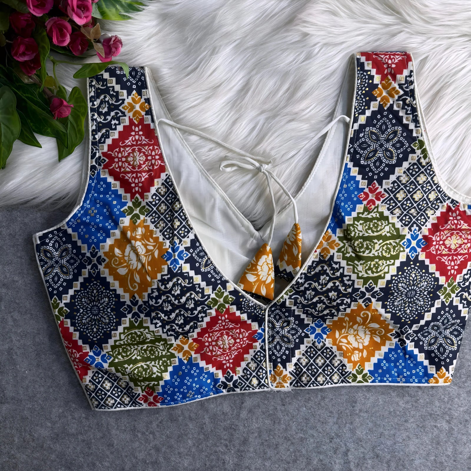Traditional blouse featuring a multicolor patchwork pattern with intricate floral and geometric motifs, deep V-neckline, white silk side panels with gold accents, and patterned tasseled tie-back detail, displayed flat on a textured grey surface with artificial pink flowers and a white fur background.