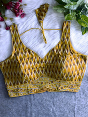 Mustard yellow silk blouse featuring traditional Ikat-inspired geometric print in brown and cream, wide shoulder straps, structured cups, and a decorative floral patterned border at the hem. Includes a matching tie-back detail with a petal-shaped tassel, displayed flat on a white fur background with pink artificial flowers and green foliage at the corners.