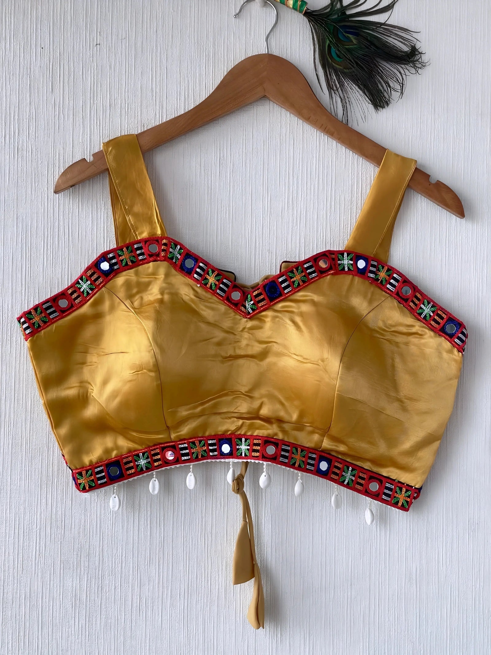 Mustard yellow ethnic blouse featuring colorful mirror work and embroidered geometric border in red, blue, and green, adorned with pearl drop tassels and a tie-up at the back, displayed on a wooden hanger against a textured off-white wall, creating a festive and traditional look.
