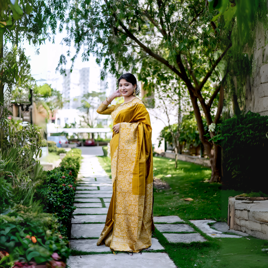 A woman models a luxurious Golden Yellow Raw Silk Saree. The saree features a wide border and pallu with dense, tone-on-tone gold Zari weaving in a Kalamkari-style floral and paisley pattern. She wears a matching long-sleeve yellow blouse and traditional gold jewelry, including a maang tikka, posing outdoors on a sunlit garden path.
