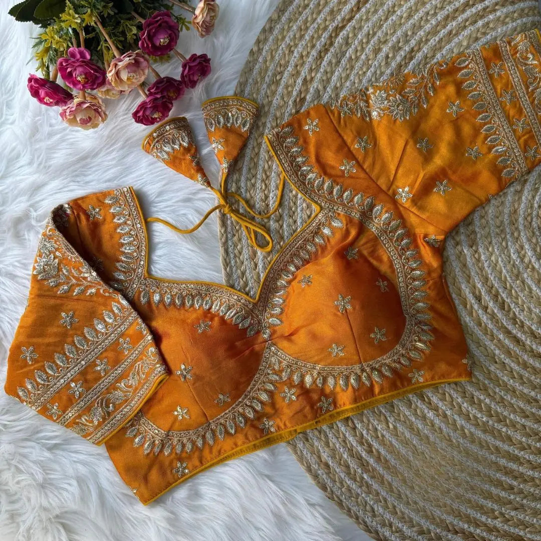 Mustard yellow blouse with intricate silver zari embroidery and floral motifs, featuring decorative borders and tie-up tassels, displayed on a textured rug with pink flowers nearby