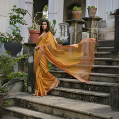 A woman models a stunning Golden Yellow Soft Silk Saree. The saree features subtle metallic motifs on the body and a wide, contrasting Copper/Rust Zari woven border. The lightweight fabric flows elegantly as she poses on a weathered stone staircase, wearing a delicate gold choker necklace.