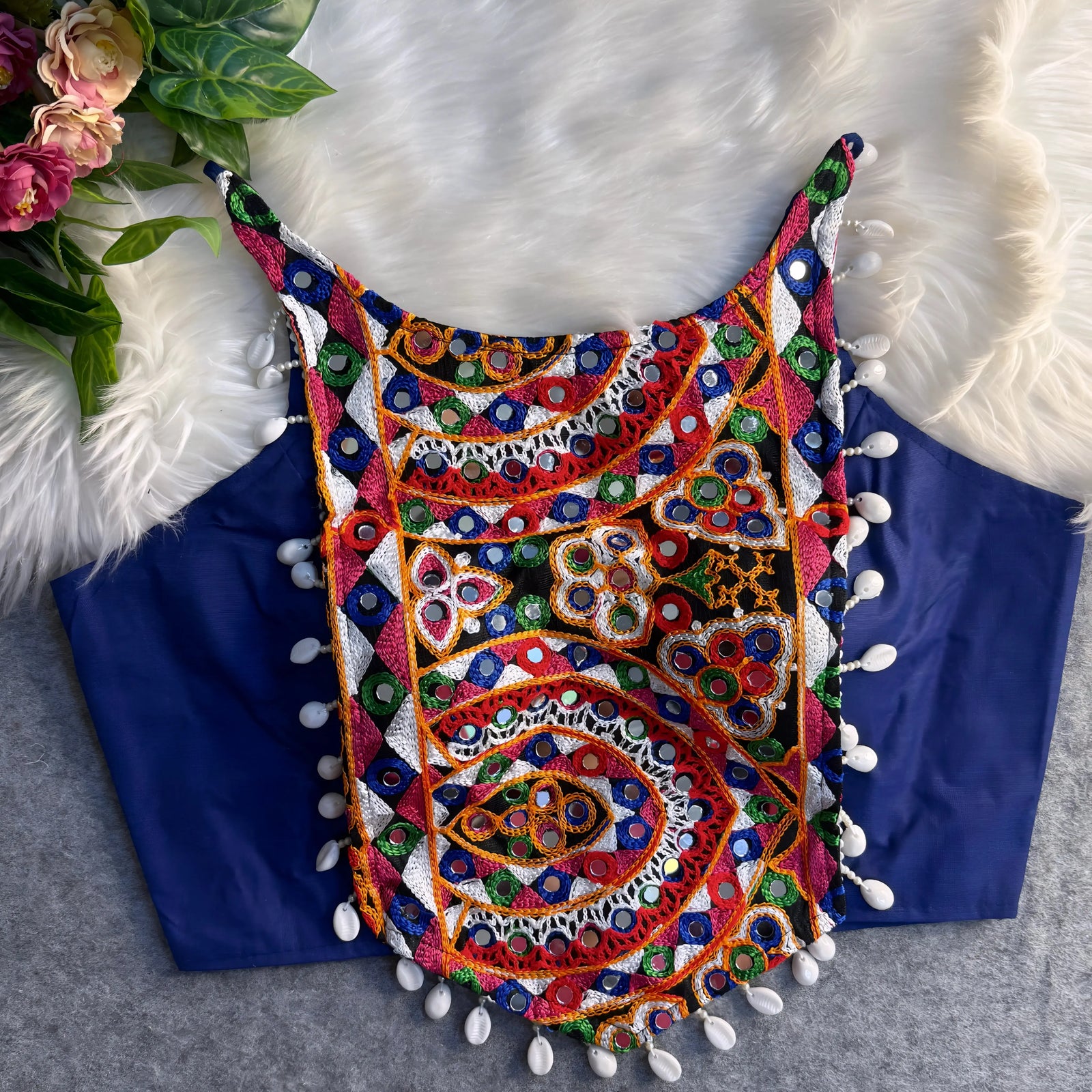 Traditional navy blue blouse featuring vibrant multicolor mirror work and intricate thread embroidery in geometric and paisley patterns, accented with white shell tassels along the hem, displayed flat on a soft fur and grey background with floral and leafy decor.