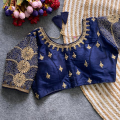 Traditional navy blue silk blouse featuring intricate gold embroidery and beadwork with floral and paisley motifs on the sleeves and neckline, displayed flat on a textured surface with a bunch of artificial flowers and a woven mat in the background.