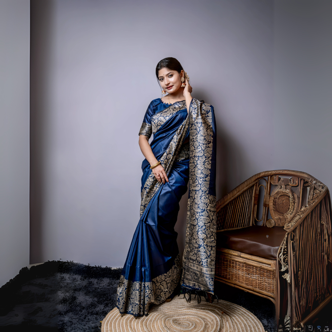A woman models a luxurious Navy Blue Raw Silk Saree. The saree features a wide border and pallu with dense, intricate gold Zari weaving in a Kalamkari-style floral and paisley pattern. She wears a matching navy blouse and traditional gold jewelry, including large jhumkas and gold bangles, with white flowers adorning her hair.