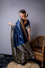A woman models a luxurious Navy Blue Raw Silk Saree. The saree features a wide border and pallu with dense, intricate gold Zari weaving in a Kalamkari-style floral and paisley pattern. She wears a matching navy blouse and traditional gold jewelry, including large jhumkas and gold bangles, with white flowers adorning her hair.