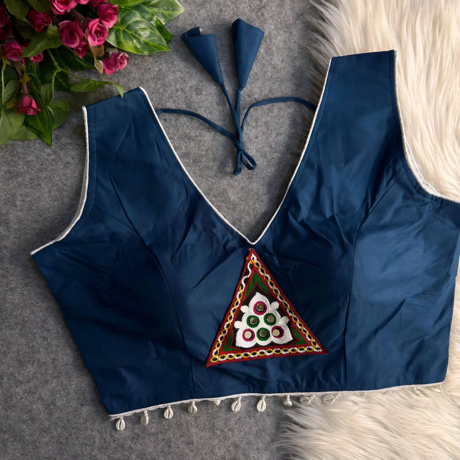 Navy blue padded blouse with white piping, featuring a central embroidered triangle motif with mirror work and shell detailing along the hem, displayed on a grey surface with artificial pink flowers and white fur in the background.